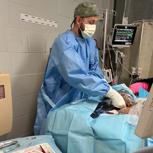 Resident wearing sterile gown, gloves, mask, and surgical cap performing a bedside procedure on a patient in a hospital room while monitoring equipment displays vital signs.