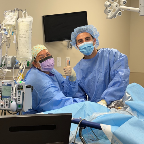 Two residents in sterile gowns, masks, and caps performing a medical procedure at a patient's bedside in a hospital treatment room.