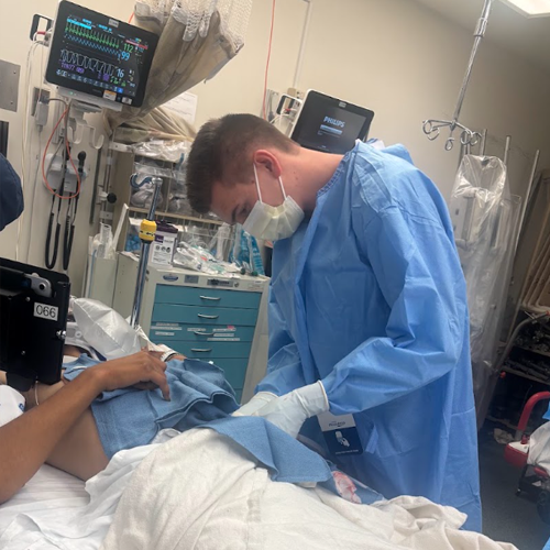 Resident in sterile protective attire performing a bedside medical procedure on a patient in a hospital room with monitoring equipment visible in the background