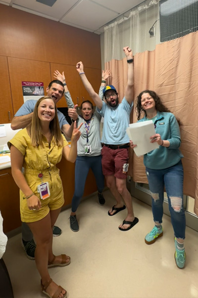 Five fellows wearing ID badges pose in a hospital exam room, raising their arms and giving thumbs-up gestures