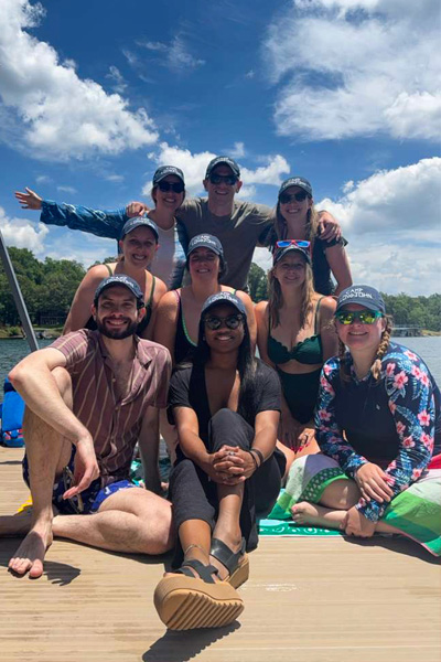Nine fellows pose together on a dock beside a lake under a bright blue sky with scattered clouds
