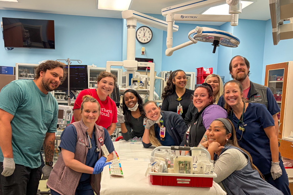 Pediatric Emergency Medicine fellows gathered around a simulation bed in a hospital training room.