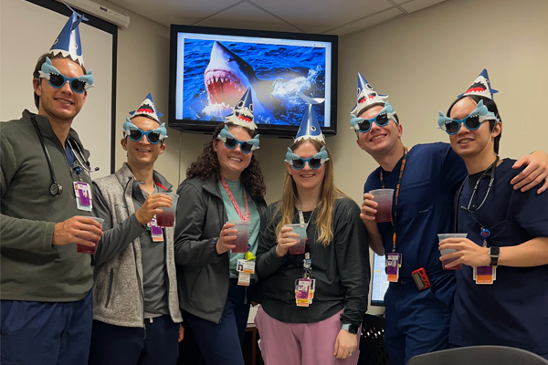 Six residents on party hats and sunglasses with a shark on a TV behind them