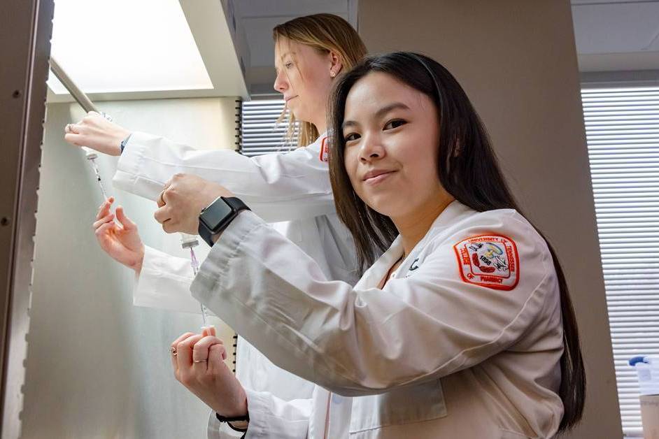 Pharmacy students hold syringe and bottle in white lab coats.